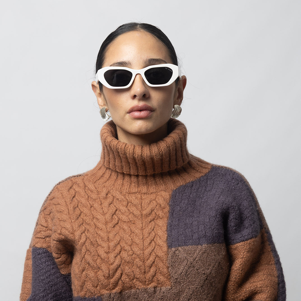 Person wearing a brown and purple patterned sweater with white sunglasses on a light gray background