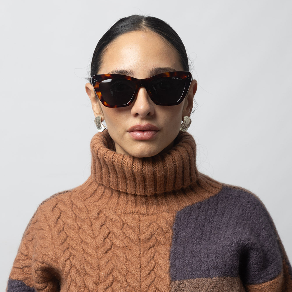 Woman wearing a brown and purple sweater with sunglasses against a light gray background