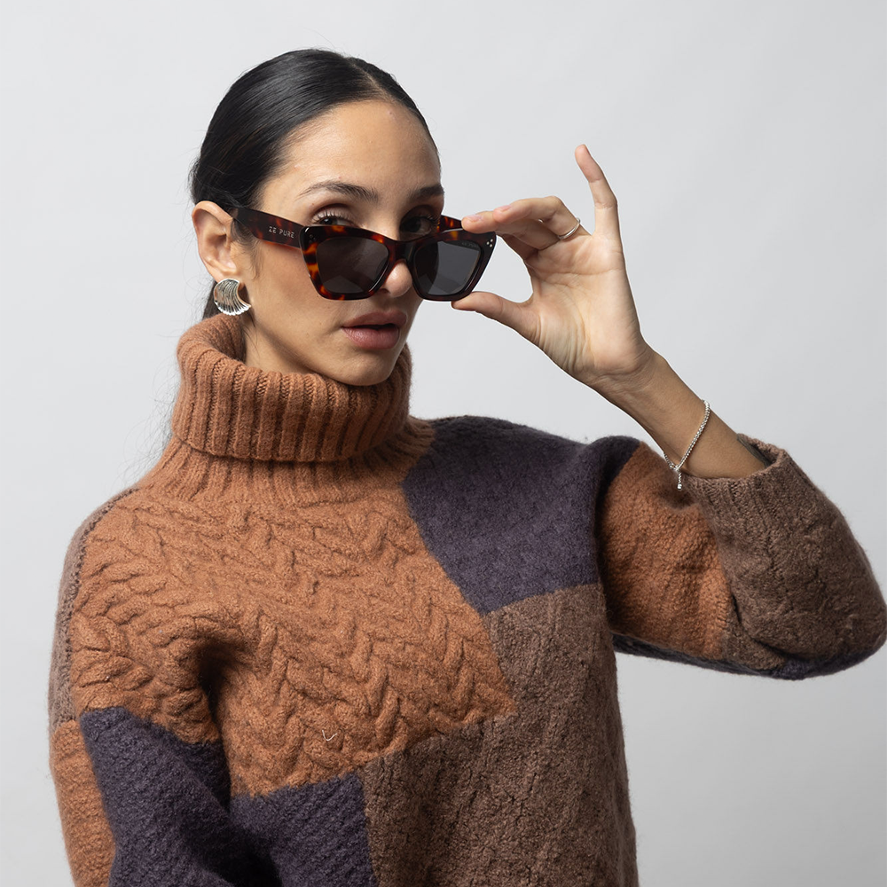 Woman wearing a brown and purple sweater with sunglasses, against a plain background