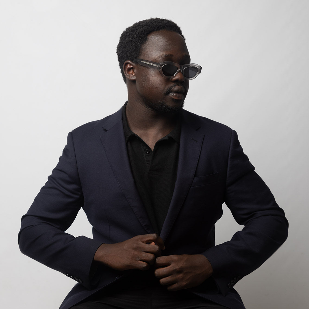Man wearing a dark suit and sunglasses against a plain background