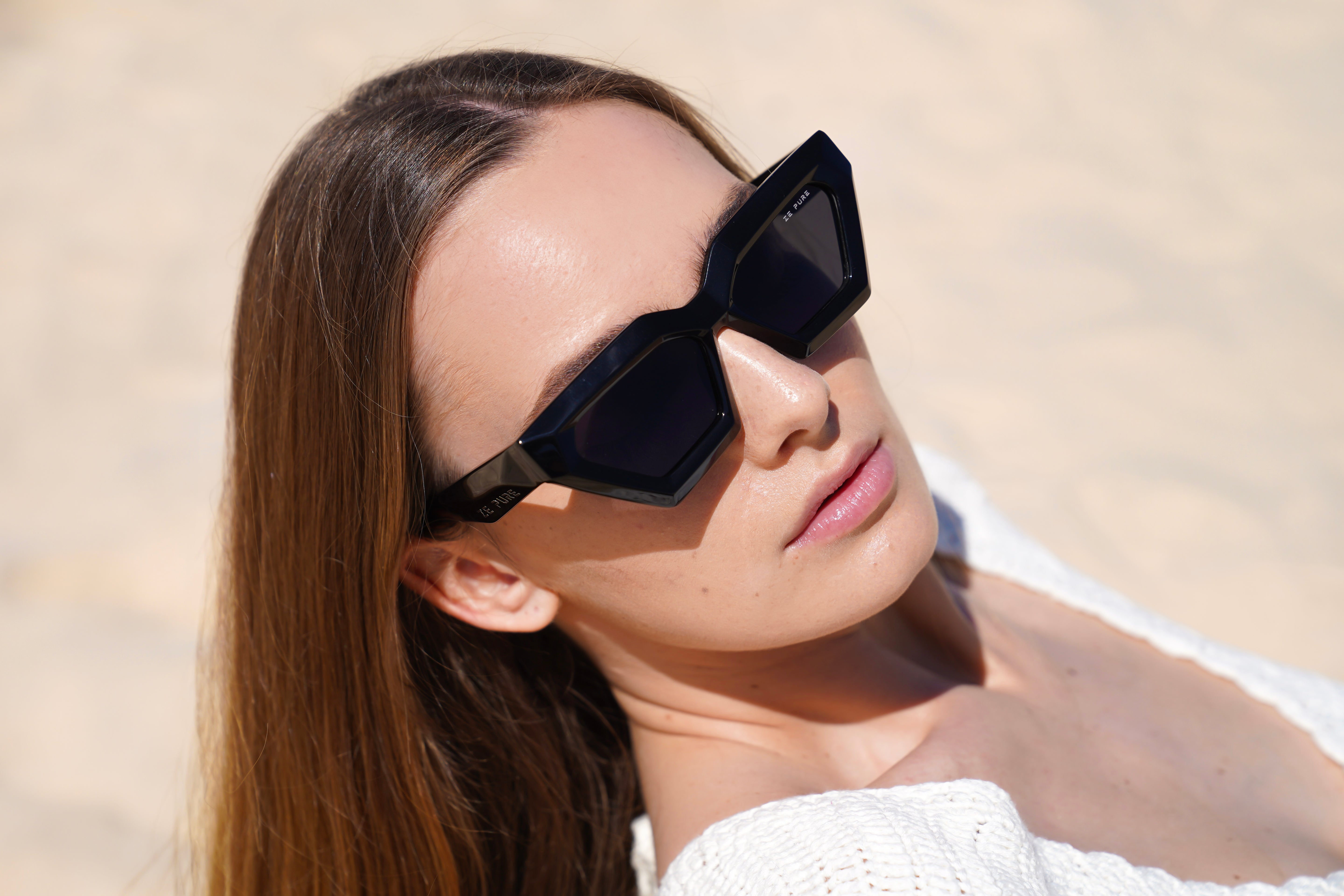 A person lying on a sandy surface wearing black sunglasses and a white sweater.